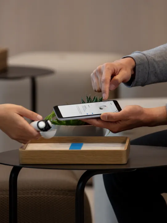 A hand holds Porcelain-colored Pixel Buds over a dark brown side table. Another hand points towards a phone screen.