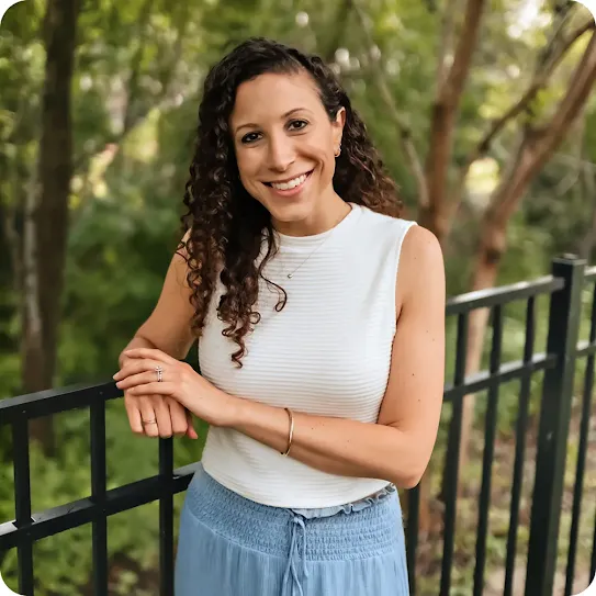 A smiling woman with long, curly dark hair leans against a black metal fence. She is wearing a white sleeveless knit top and a light blue skirt, with a soft-focus background of green trees and foliage.